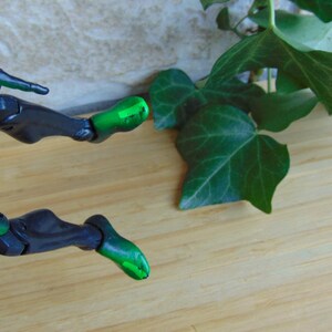 May include: A close-up of a green and black action figure's legs, with articulated joints, positioned on a light brown wooden surface. Green leaves are in the background.