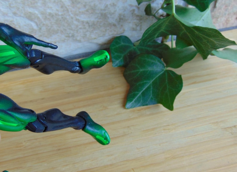 May include: A close-up of a green and black action figure's legs, with articulated joints, positioned on a light brown wooden surface. Green leaves are in the background.