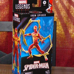 Pu&ograve; includere: Scatola della action figure Marvel Legends Iron Spider. La scatola nera presenta dettagli rossi e gialli, con immagini del personaggio Iron Spider. Il logo Marvel Spider-Man e un avvertimento sulle piccole parti sono anch'essi presenti.