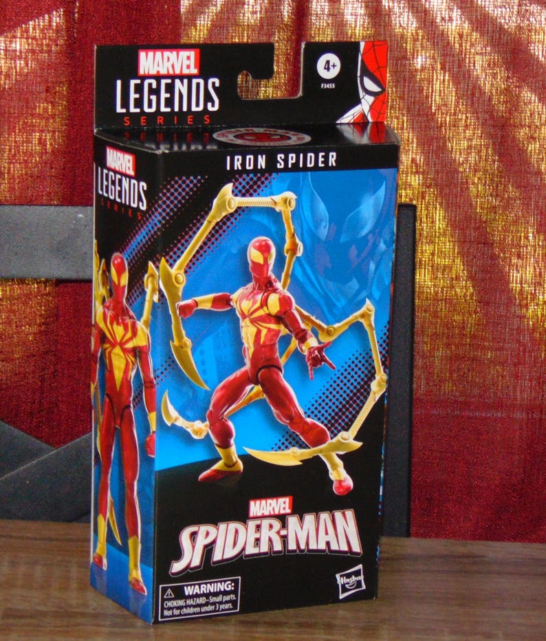 Pu&ograve; includere: Scatola della action figure Marvel Legends Iron Spider. La scatola nera presenta dettagli rossi e gialli, con immagini del personaggio Iron Spider. Il logo Marvel Spider-Man e un avvertimento sulle piccole parti sono anch'essi presenti.