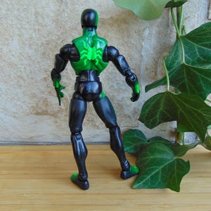 May include: A black and green action figure of a superhero, viewed from the back. The figure has a green spider emblem on its back and green accents on the hands and feet. The figure is posed against a neutral background.