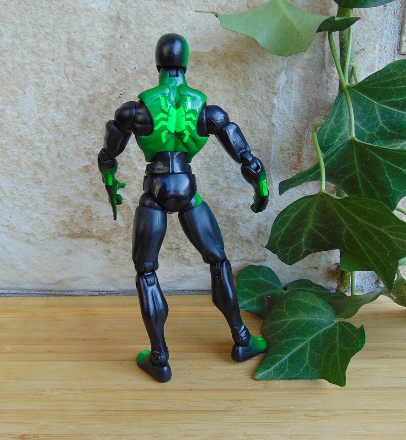 May include: A black and green action figure of a superhero, viewed from the back. The figure has a green spider emblem on its back and green accents on the hands and feet. The figure is posed against a neutral background.