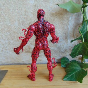 May include: A red action figure with black vein-like patterns. The figure has a muscular build and is posed with arms outstretched. The figure has tendrils extending from the arms and legs. The figure is standing on a wooden surface.