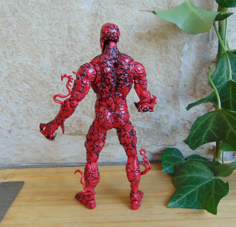 May include: A red action figure with black vein-like patterns. The figure has a muscular build and is posed with arms outstretched. The figure has tendrils extending from the arms and legs. The figure is standing on a wooden surface.