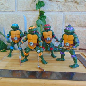 May include: Four green turtle action figures with orange chest plates, each wearing a different colored mask and belt with a lettered buckle. They hold various weapons including swords, nunchucks, and a bo staff. A metal ruler is visible in the foreground.