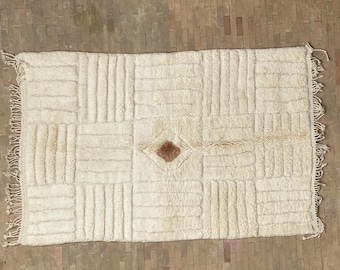 May include: A white, fluffy rug with a geometric pattern and a brown diamond in the center. The rug has fringe on all sides.