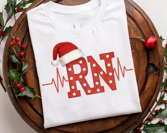 May include: A white t-shirt featuring a red graphic of the letters RN with white polka dots. The letters are topped with a red and white Santa hat and are intersected by a red EKG heart rhythm line that extends horizontally across the shirt.
