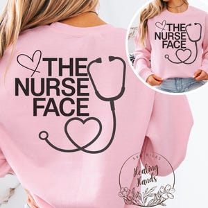 以下が含まれることがあります： 「THE NURSE FACE」の文字と黒い聴診器のグラフィックがプリントされた、薄いピンク色のスウェットシャツ。デザインにはハートの形が含まれており、スウェットシャツの背面に表示されています。スウェットシャツの小さな画像が右上隅にあります。