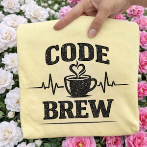May include: A light yellow t-shirt with the words "CODE BREW" in a distressed black font. A coffee cup with a heart-shaped steam design is centered between the words, with a heartbeat line extending from each side.