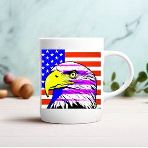 May include: White ceramic mug featuring a stylized image of an American bald eagle with a yellow beak and purple and blue accents. The background includes the American flag. The mug has a white handle and is suitable for hot or cold beverages.