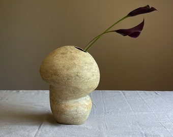 May include: A beige, mushroom-shaped ceramic vase with a textured surface. Two dark purple calla lilies emerge from the vase. The vase sits on a light gray linen surface against a neutral background.