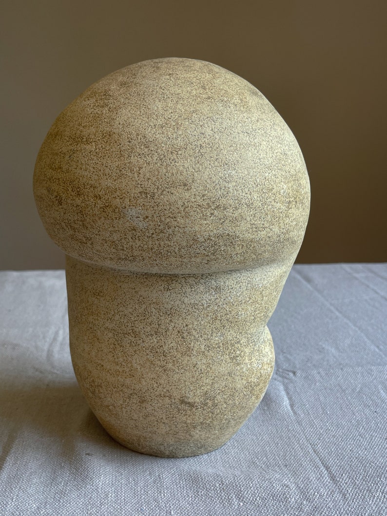 Puede incluir: Una escultura beige con forma de hongo y superficie texturizada. La escultura tiene una parte superior redondeada y una base bulbosa, creando una forma org&aacute;nica &uacute;nica. El color neutro y el dise&ntilde;o sencillo la convierten en una pieza decorativa vers&aacute;til.