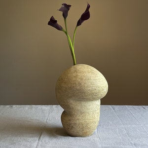 Puede incluir: Un jarr&oacute;n de cer&aacute;mica beige con forma org&aacute;nica que contiene tres calas moradas oscuras. El jarr&oacute;n tiene una superficie texturizada y una forma redondeada &uacute;nica. Las flores tienen tallos verdes, creando un arreglo floral simple y elegante.