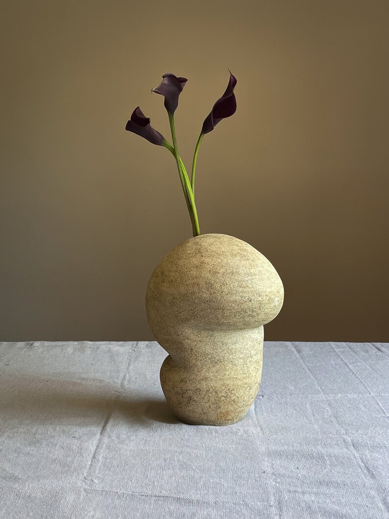 Puede incluir: Un jarr&oacute;n de cer&aacute;mica beige con forma org&aacute;nica que contiene tres calas moradas oscuras. El jarr&oacute;n tiene una superficie texturizada y una forma redondeada &uacute;nica. Las flores tienen tallos verdes, creando un arreglo floral simple y elegante.