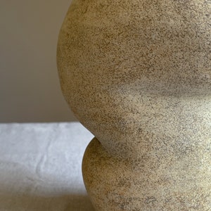 Puede incluir: Un jarr&oacute;n de cer&aacute;mica beige texturizado con una forma bulbosa &uacute;nica. El jarr&oacute;n tiene una superficie rugosa y moteada y un dise&ntilde;o de dos partes, con una esfera m&aacute;s peque&ntilde;a conectada a una m&aacute;s grande. El color neutro y la forma org&aacute;nica sugieren una est&eacute;tica moderna y minimalista.
