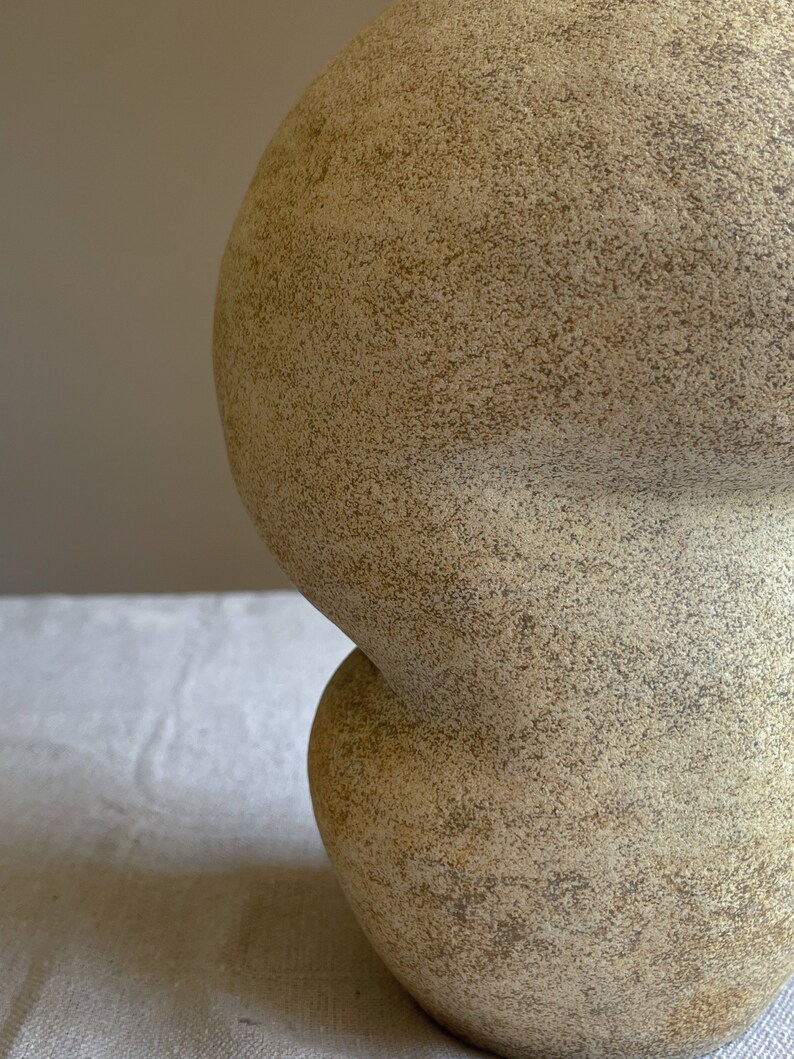 Puede incluir: Un jarr&oacute;n de cer&aacute;mica beige texturizado con una forma bulbosa &uacute;nica. El jarr&oacute;n tiene una superficie rugosa y moteada y un dise&ntilde;o de dos partes, con una esfera m&aacute;s peque&ntilde;a conectada a una m&aacute;s grande. El color neutro y la forma org&aacute;nica sugieren una est&eacute;tica moderna y minimalista.
