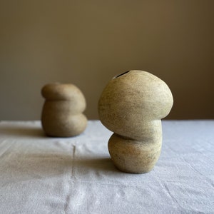Puede incluir: Dos jarrones de cer&aacute;mica beige con superficie texturizada. Los jarrones tienen una forma org&aacute;nica &uacute;nica, con una base bulbosa y una parte superior redondeada. Un jarr&oacute;n est&aacute; en primer plano y el otro est&aacute; ligeramente borroso en el fondo. Los jarrones est&aacute;n sobre un mantel de lino blanco.
