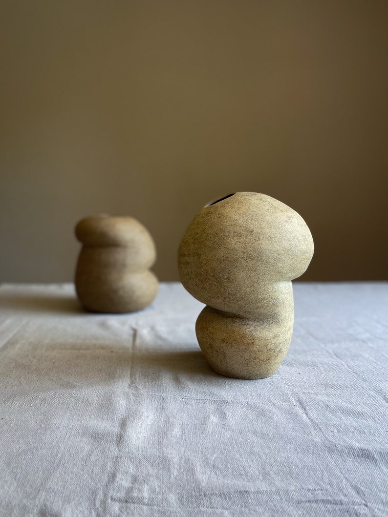 Puede incluir: Dos jarrones de cer&aacute;mica beige con superficie texturizada. Los jarrones tienen una forma org&aacute;nica &uacute;nica, con una base bulbosa y una parte superior redondeada. Un jarr&oacute;n est&aacute; en primer plano y el otro est&aacute; ligeramente borroso en el fondo. Los jarrones est&aacute;n sobre un mantel de lino blanco.