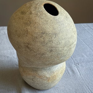 Puede incluir: Un jarr&oacute;n de cer&aacute;mica beige con una forma org&aacute;nica &uacute;nica. El jarr&oacute;n tiene una parte superior redondeada con una gran abertura y una base bulbosa. La superficie tiene un aspecto texturizado y moteado con tonos grises y marrones m&aacute;s oscuros. Es un art&iacute;culo decorativo.