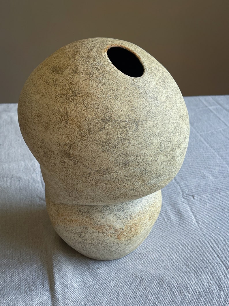 Puede incluir: Un jarr&oacute;n de cer&aacute;mica beige con una forma org&aacute;nica &uacute;nica. El jarr&oacute;n tiene una parte superior redondeada con una gran abertura y una base bulbosa. La superficie tiene un aspecto texturizado y moteado con tonos grises y marrones m&aacute;s oscuros. Es un art&iacute;culo decorativo.