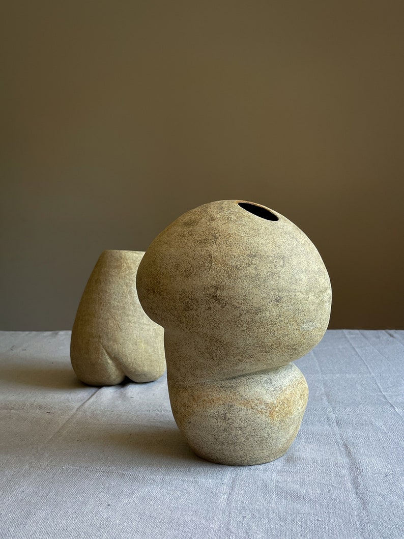Puede incluir: Dos jarrones de cer&aacute;mica beige con formas org&aacute;nicas. El jarr&oacute;n en primer plano tiene una parte superior bulbosa con una abertura oscura y una base redondeada. El segundo jarr&oacute;n tiene una forma m&aacute;s alargada. Ambos jarrones tienen una superficie texturizada.