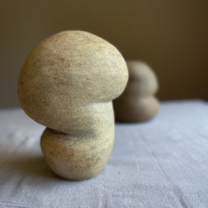 Puede incluir: Una escultura de cer&aacute;mica beige con forma de hongo y superficie texturizada. La escultura tiene un sombrero redondeado y un tallo curvo, y se asienta sobre una superficie de lino gris claro. Otra escultura similar est&aacute; borrosa en el fondo.