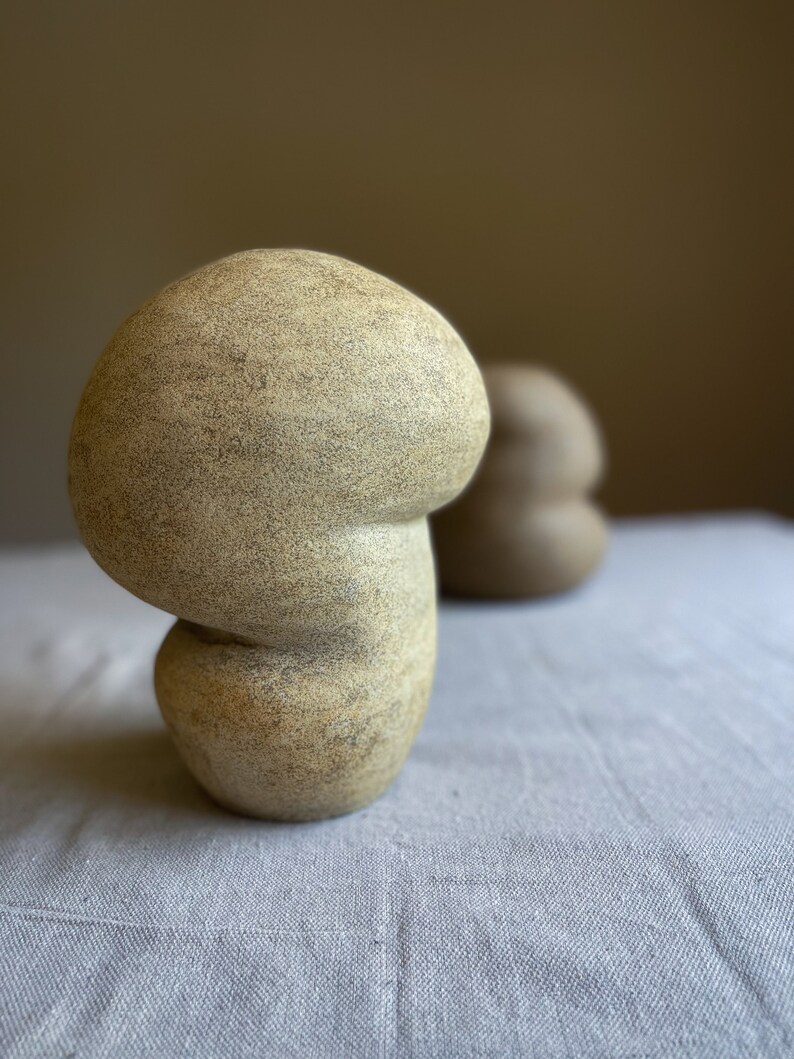 Puede incluir: Una escultura de cer&aacute;mica beige con forma de hongo y superficie texturizada. La escultura tiene un sombrero redondeado y un tallo curvo, y se asienta sobre una superficie de lino gris claro. Otra escultura similar est&aacute; borrosa en el fondo.