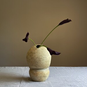 Puede incluir: Un jarr&oacute;n de cer&aacute;mica beige texturizado con forma bulbosa, que contiene dos calas moradas oscuras. El jarr&oacute;n tiene un dise&ntilde;o &uacute;nico en capas y est&aacute; sobre una superficie de lino blanco. El fondo es de color neutro.