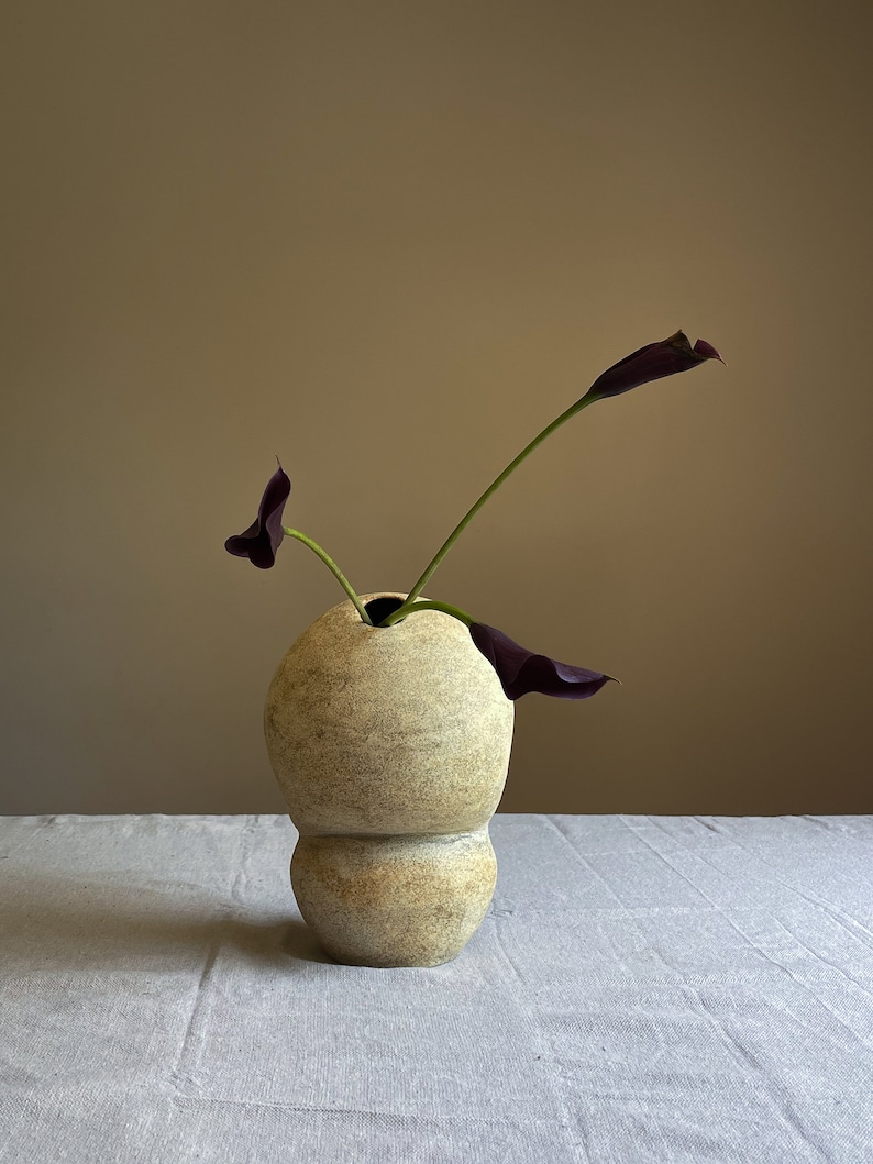Puede incluir: Un jarr&oacute;n de cer&aacute;mica beige texturizado con forma bulbosa, que contiene dos calas moradas oscuras. El jarr&oacute;n tiene un dise&ntilde;o &uacute;nico en capas y est&aacute; sobre una superficie de lino blanco. El fondo es de color neutro.