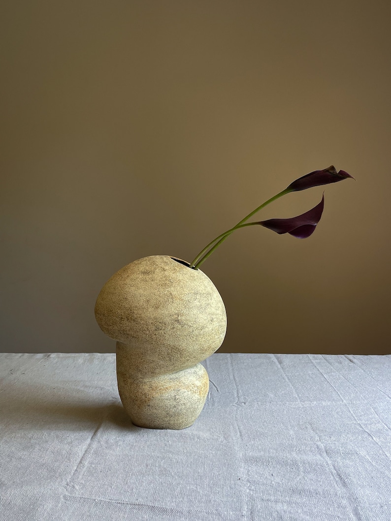 Puede incluir: Un jarr&oacute;n de cer&aacute;mica beige con forma de hongo y superficie texturizada. Dos calas moradas oscuras emergen del jarr&oacute;n. El jarr&oacute;n est&aacute; sobre una superficie de lino gris claro, sobre un fondo neutro.