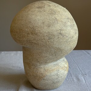 Puede incluir: Una escultura beige en forma de hongo con una superficie texturizada. La escultura tiene un sombrero redondeado y un tallo bulboso, creando una forma org&aacute;nica caprichosa. El color neutro y la forma &uacute;nica lo convierten en una pieza decorativa.