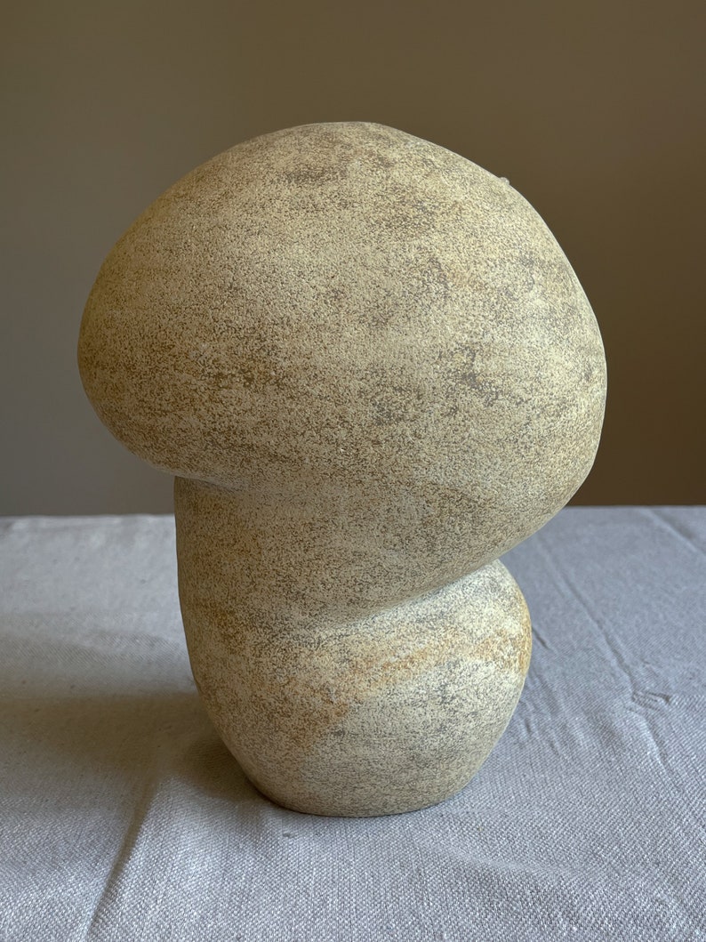 Puede incluir: Una escultura beige en forma de hongo con una superficie texturizada. La escultura tiene un sombrero redondeado y un tallo bulboso, creando una forma org&aacute;nica caprichosa. El color neutro y la forma &uacute;nica lo convierten en una pieza decorativa.