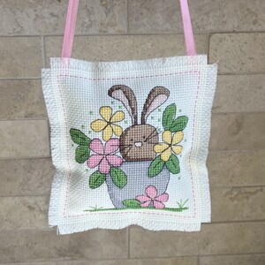 May include: A white cross-stitch with a brown bunny holding a bouquet of yellow, pink, and green flowers in a gray pot. The cross-stitch is hanging from a pink ribbon.