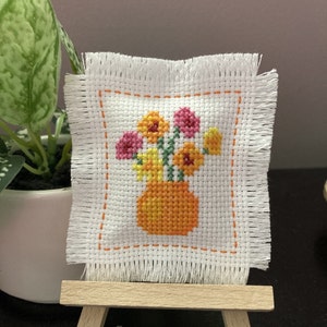 May include: A small, square cross-stitch embroidery featuring a vase of colorful flowers. The vase is orange, and the flowers are pink, yellow, and orange. The embroidery is on white fabric with a fringed edge and an orange stitched border. It is displayed on a miniature wooden easel.