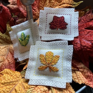 May include: Three small, square, off-white fabric patches with embroidered leaf designs in shades of green, red, and orange. The patches are surrounded by fall-colored leaves, creating a seasonal theme.