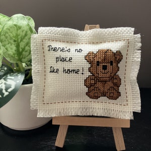 May include: A white cross-stitch with a brown teddy bear and the text "There's no place like home!" displayed on a small wooden easel.