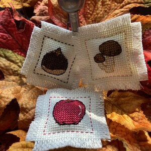 May include: Hand-stitched miniature cross-stitch art featuring a chestnut, mushrooms, and a red apple. The artwork is displayed on small, square pieces of off-white fabric with a stitched border. The background includes fall leaves.