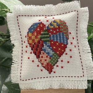 May include: A small white pillow with a red fringe border, featuring a colorful patchwork heart design. The pillow is displayed on a small wooden easel.