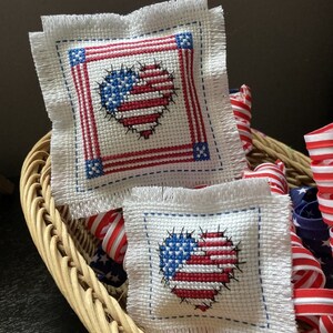 May include: Two small white fabric squares with fringed edges, each embroidered with a red, white, and blue heart-shaped American flag. The squares are in a wicker basket with red, white, and blue ribbon.