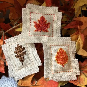 May include: Three small, square, off-white fabric patches with embroidered fall leaf designs. The leaves are in shades of orange and brown, with red stitching around the edges. The patches are set against a backdrop of colorful autumn leaves.