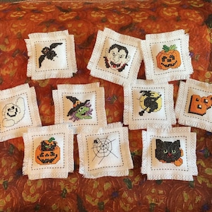 May include: A collection of square, white fabric patches with frayed edges, each featuring a cross-stitched Halloween design. Designs include a bat, vampire, jack-o'-lantern, ghost, witch, moon, the word "BOO", spider web, and a black cat.