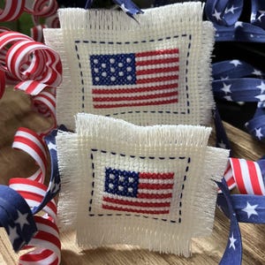 May include: Two small, square, off-white fabric ornaments with cross-stitched American flags. The flags feature red and white stripes and a blue field with white stars. Decorative red, white, and blue ribbon and curly ribbon are in the background.
