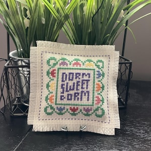 May include: Square cross-stitch with the words "DORM SWEET DORM" in purple, green, and yellow. The cross-stitch is framed with a green border and colorful floral accents. The background includes a wire basket and artificial plants.