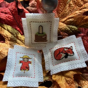 May include: Three small, off-white cross-stitch patches with autumn-themed designs: a basket, a scarecrow, and a sleeping fox. The patches are displayed on a bed of colorful fall leaves, with a silver knob in the background.