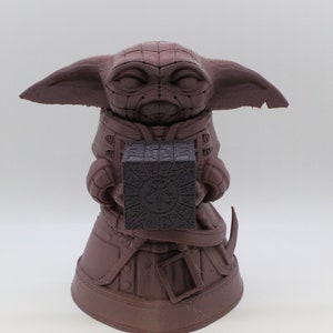 May include: A brown 3D printed figurine of a character holding a black cube. The character has large ears and a hooded robe.