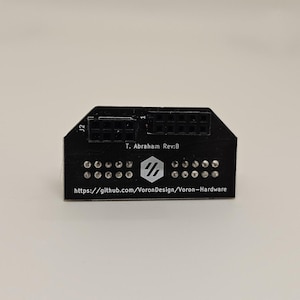 May include: A black circuit board with a connector, labeled "J2", and a small white logo with the text "T. Abraham Rev:B" and a website address: "https://github.com/VoronDesign/Voron-Hardware".