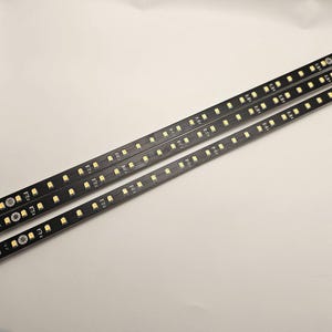 Op de afbeelding: Drie zwarte LED-lichtstrips met gelijkmatig verdeelde, kleine, gele lampjes. De strips zijn diagonaal over een wit oppervlak geplaatst. De lampjes hebben een warme gele kleur.