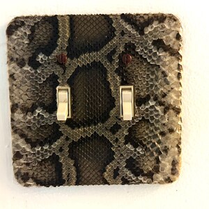 May include: A decorative light switch cover featuring a snakeskin pattern. The cover has two light switches with off-white toggles and two small red screws. The snakeskin pattern is a mix of gray, brown, and black.