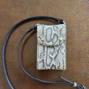 May include: A rectangular, snakeskin-patterned crossbody phone bag with a black leather strap. The bag has a flap closure secured by two gold-colored rivets. The strap is adjustable and features a silver-colored buckle.