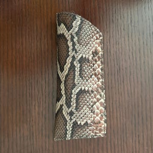 May include: A brown and tan snakeskin-patterned eyeglass case. The case is rectangular with a curved top edge. The pattern features a mix of brown, tan, and cream-colored scales, creating a realistic snakeskin appearance. The case is closed.